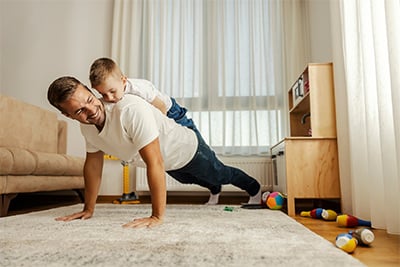 A strong father is doing pushups at home with his son on his backs.